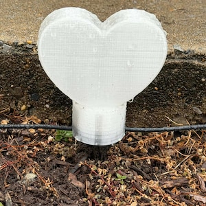 May include: A white, heart-shaped garden stake. The stake is made of a textured material and is placed in the ground. The heart is approximately 4 inches wide and 3 inches tall.