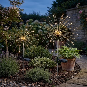 May include: Two solar-powered garden lights with a starburst design. Each light has a central sphere with numerous thin, flexible wires radiating outwards, each tipped with a warm white LED. The lights are set in a garden with plants and a stone wall.