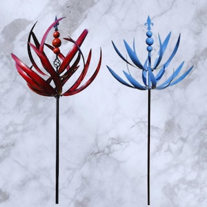 Puede incluir: Dos molinillos de jardín de metal, uno rojo y otro azul, cada uno con un tallo central y aspas curvas en forma de pétalo. El molinillo rojo tiene una cuenta decorativa y un elemento en espiral, mientras que el azul presenta un diseño de flor de lis. Decoraciones de jardín.