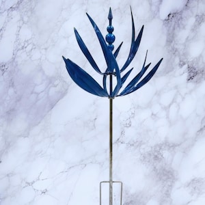 Puede incluir: Una estaca de jardín de metal azul con un diseño floral decorativo. La parte superior presenta formas curvas en forma de hojas y tres acentos esféricos. La estaca es de metal y tiene una base simple de tres puntas para una fácil colocación en el jardín.