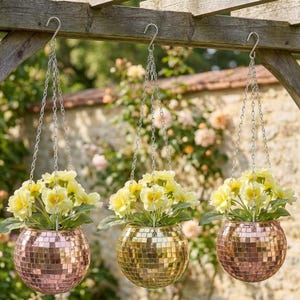 May include: Three hanging planters with yellow flowers. The spherical planters are covered in small, reflective tiles in shades of gold and rose gold. Each planter hangs from a silver chain attached to a wooden beam.