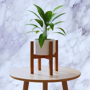 Op de afbeelding: Een kamerplant met lange groene bladeren in een witte keramische pot. De pot staat op een plantenstandaard van natuurlijk hout. De standaard staat op een ronde houten tafel. De achtergrond is een marmerpatroon.