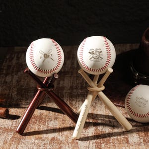 May include: Two white baseballs with red stitching, each resting on a tripod stand made of three miniature wooden baseball bats. One stand is dark brown, the other is light tan. The baseballs feature custom engravings, including text and graphics.