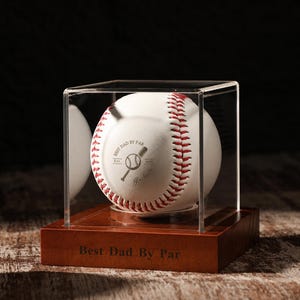 Könnte beinhalten: Ein weißer Baseball mit roten Nähten in einer durchsichtigen Acryl-Vitrine auf einem Holzsockel. Der Ball zeigt eine Grafik mit dem Text Best Dad By Par, und der Holzsockel ist mit demselben Text, Best Dad By Par, graviert.