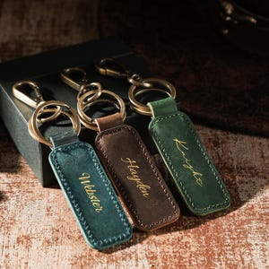May include: Three rectangular leather keychains with antique brass hardware. The keychains are teal, dark brown, and forest green, each featuring gold-colored cursive text. The teal keychain reads Webster, the brown reads Hayden, and the green reads Knight.
