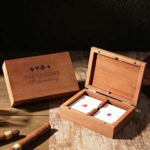 May include: A wooden playing card box with the text "THE TAYLORS 12th Anniversary" engraved on the lid. The box is open, revealing two playing cards. A glass of amber liquid and two cigars are also visible.