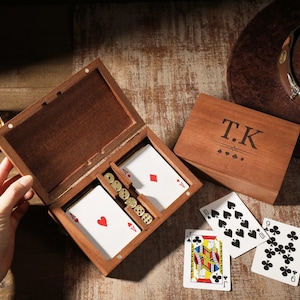 May include: A wooden card game set with a deck of cards, dice, and poker chips. The box is open, revealing the contents. A brown cowboy hat and a glass of whiskey are in the background. The box lid is engraved with the initials "T.K."