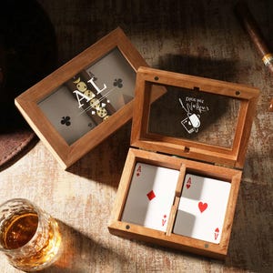 Puede incluir: Caja de madera para naipes con tapa de cristal, que muestra dos Ases. La caja está abierta, revelando las cartas. La caja superior tiene las iniciales "AL" y un diseño de naipe. Un vaso de líquido ámbar y cigarros también están en la imagen.