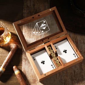 May include: Wooden playing card box with a glass top, containing playing cards and poker chips. Two cigars and a glass of amber liquid sit next to the box. The box has a clear top with a playing card design.