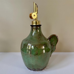 Op de afbeelding: Een keramische oliekruik met een gespikkelde groene en bruine glazuurlaag, voorzien van een ronde vorm, een zijhandvat en een goudkleurige metalen schenktuit met een scharnierend deksel. Het object heeft een gladde, glanzende afwerking.