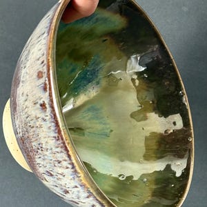 Op de afbeelding: Een handgemaakte keramische kom met een uniek glazuur. De binnenkant heeft tinten groen en blauw, terwijl de buitenkant een gespikkelde, getextureerde uitstraling heeft met bruine en crèmetinten. De rand is donkerbruin. De kom heeft een kleine, lichtgekleurde basis.