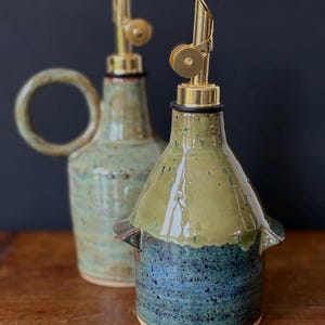 Op de afbeelding: Twee keramische olieflessen met messing schenktuiten. De flessen zijn geglazuurd in groen- en blauwtinten, met een gespikkelde textuur. Eén fles heeft een handvat, de andere kleine zijhandvatten. De schenktuiten zijn goudkleurig en hebben een hendelmechanisme.