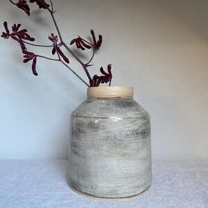Puede incluir: Un jarrón de cerámica con cuerpo cilíndrico y borde beige claro. El jarrón tiene un acabado texturizado, moteado en gris y blanco. Una ramita de flores de color burdeos oscuro se extiende desde el jarrón, creando una estética natural y minimalista.