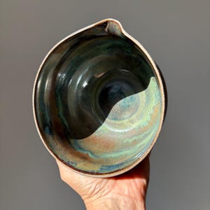 Pode incluir: Uma tigela de cerâmica com formato arredondado e um bico vertedor. O interior apresenta uma mistura de esmaltes preto profundo, azul escuro e verde terroso que se misturam, criando um acabamento liso e brilhante com variações de cor.