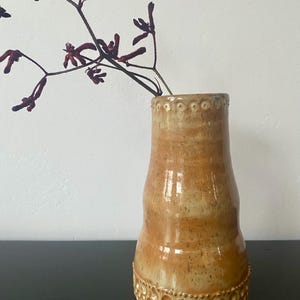 Può includere: Un vaso in ceramica fatto a mano, dai toni caldi e terrosi. Il vaso presenta un motivo strutturato a forma di bolle alla base e piccole rientranze circolari vicino al bordo. Steli e boccioli rosso scuro sono posti all'interno del vaso.