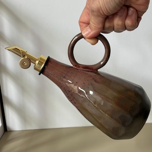 Op de afbeelding: Een bruine keramische oliedispenser met een gefacetteerd, veelzijdig lichaam en een cirkelvormig handvat. De houder heeft een glanzend, gevlekt bruin glazuur en is voorzien van een goudkleurige metalen schenktuit met een zwarte rubberen ring.