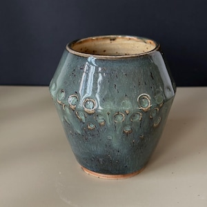 Op de afbeelding: Een handgemaakte keramische vaas met een turquoise glazuur en een textuurpatroon van cirkelvormige inkepingen. De vaas heeft een brede, facetgeslepen body en een lichtbruin interieur. De basis is een donkerdere bruintint.
