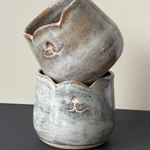 Op de afbeelding: Twee handgemaakte keramische potten op elkaar gestapeld. De potten zijn gemêleerd grijs met een geschulpte rand en een decoratief reliëf ontwerp. De potten zijn ongeveer 10 cm hoog.