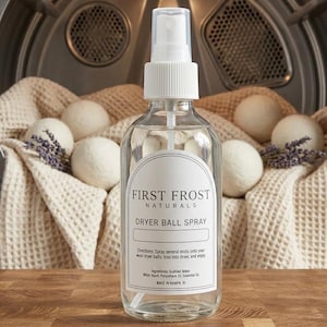 Peut inclure: Un flacon pulvérisateur en verre transparent étiqueté First Frost Naturals Dryer Ball Spray avec un embout pompe blanc. L'étiquette contient des instructions et des ingrédients. Derrière se trouvent des boules de séchage en laine et de la lavande.