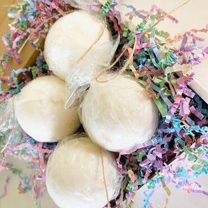 May include: Four spherical white bath bombs individually wrapped in clear plastic film, nestled within a cardboard box filled with shredded paper strips in pastel shades of pink, blue, green, and purple, tied with thin brown twine.
