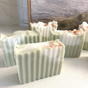 May include: Several rectangular bars of soap with a vertically ribbed texture. The soap features a two-tone design with a pale sage green base and a white top layer. Each bar is topped with small, scattered pink Himalayan salt crystals.