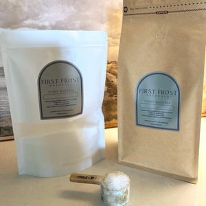May include: Two stand-up pouches of First Frost Naturals scent booster, one white and one tan, featuring arched labels with instructions. A wooden measuring scoop filled with white crystalline granules sits in the foreground, marked 1 TBSP - 2 TBSP.