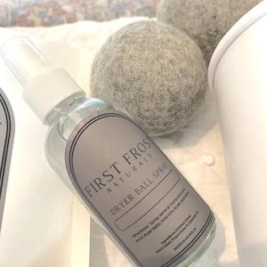 Peut inclure: Un flacon pulvérisateur en plastique transparent étiqueté First Frost Naturals Dryer Ball Spray avec un embout blanc, placé à côté de deux boules de séchage en laine grise, rondes et texturées. L'étiquette contient des instructions et ingrédients.