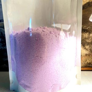 May include: A clear, stand-up plastic pouch filled with a fine, light purple powder. The container is transparent, allowing for a clear view of the granular, lavender-colored substance inside, which fills the lower portion of the upright bag.