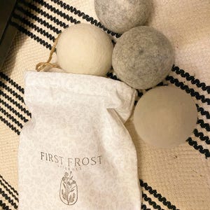 May include: A white fabric drawstring bag featuring a delicate floral pattern and the text "FIRST FROST NATURALS" above a botanical illustration. Three round, textured felt balls in shades of cream and grey are positioned near the bag.
