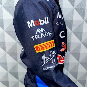 May include: Navy blue racing suit with sponsor logos including Mobil, Pirelli, and Ava Trade. The suit features a blue and red design with the words "Maui Jim" and "Polarizedplus" printed on the sleeve. The suit has a blue stripe down the side.