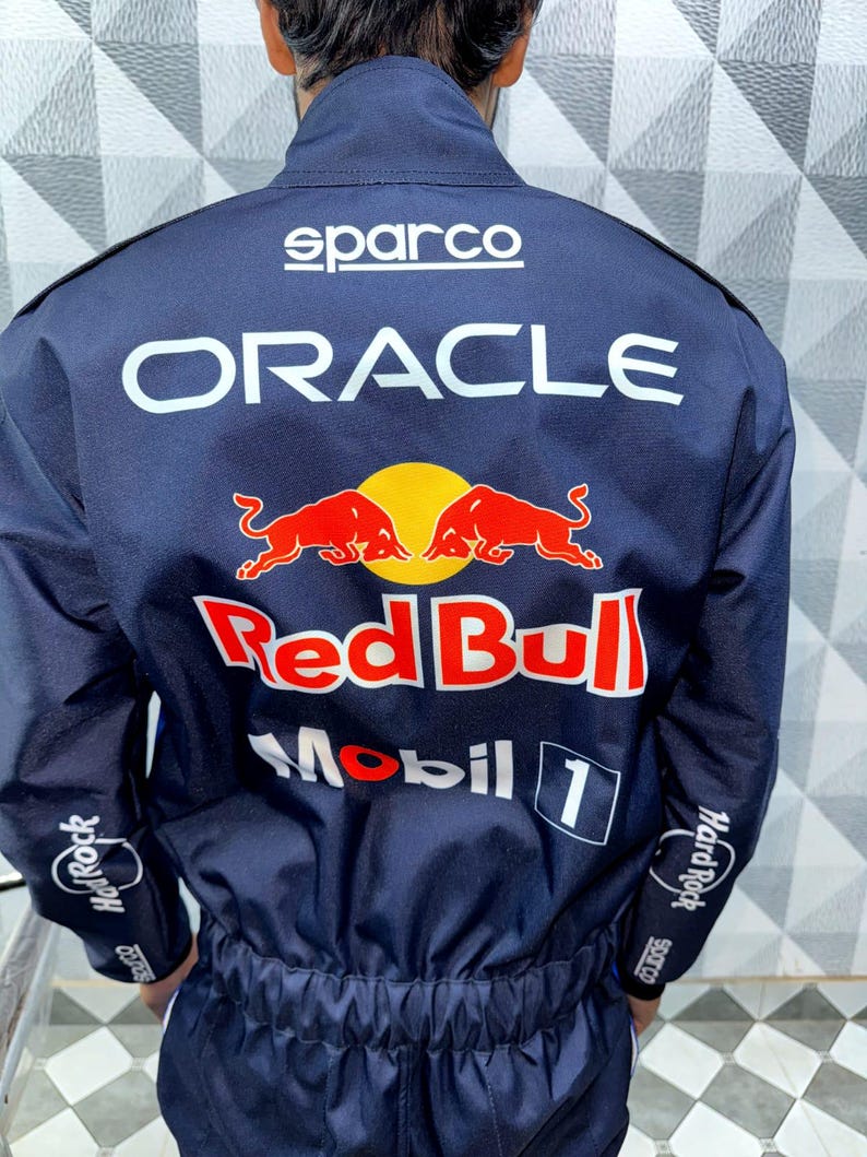 May include: Navy blue racing suit with the Sparco logo at the top and the Oracle and Red Bull logos in white and red. The sleeves have Hard Rock Cafe logos. The suit is a one-piece design.