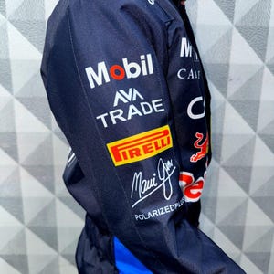 May include: Navy blue racing suit with sponsor logos including Mobil, Pirelli, and Ava Trade. The suit features a blue and red design with the words "Maui Jim" and "Polarizedplus" printed on the sleeve. The suit has a blue stripe down the side.