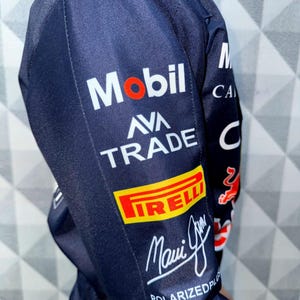 May include: Navy blue jacket with multiple sponsor logos, including "Mobil," "AVA TRADE," and "PIRELLI." The jacket features a signature and the text "POLARIZEDPLUS." The design incorporates red and white accents.