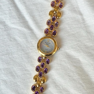 May include: A gold-toned wristwatch with a light blue face and a band adorned with alternating gold and purple oval-shaped stones. The watch face displays the brand name and the word "Quartz". The watch is set against a white fabric background.
