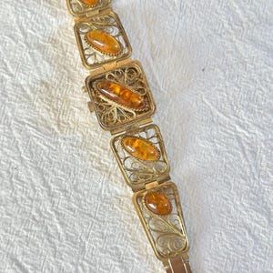 May include: A vintage gold-toned bracelet featuring rectangular filigree links, each holding an oval amber stone. The bracelet has a delicate, ornate design with a secure clasp. The amber stones have a warm, honey-like color.