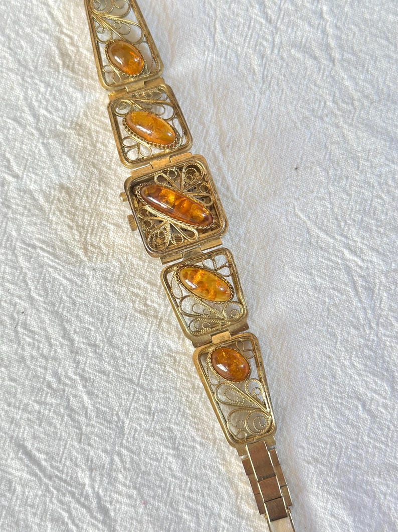May include: A vintage gold-toned bracelet featuring rectangular filigree links, each holding an oval amber stone. The bracelet has a delicate, ornate design with a secure clasp. The amber stones have a warm, honey-like color.