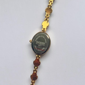May include: Gold-toned Joan Rivers Classics watch with an oval face and a linked bracelet. The watch face displays the text "Stainless Steel Back" and "Japan Movement". The bracelet features a series of small, decorative links.