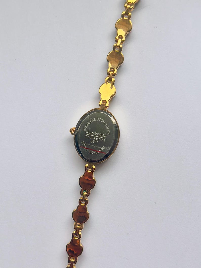 May include: Gold-toned Joan Rivers Classics watch with an oval face and a linked bracelet. The watch face displays the text "Stainless Steel Back" and "Japan Movement". The bracelet features a series of small, decorative links.