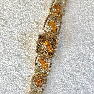May include: Vintage gold-toned bracelet featuring rectangular filigree links, each holding an amber-colored stone. The bracelet has a watch face in the center and a clasp closure. The amber stones have a warm, honey-like hue.