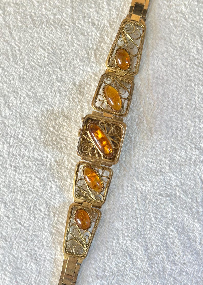 May include: Vintage gold-toned bracelet featuring rectangular filigree links, each holding an amber-colored stone. The bracelet has a watch face in the center and a clasp closure. The amber stones have a warm, honey-like hue.