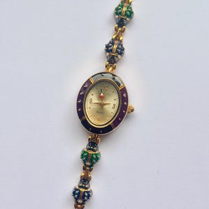 May include: Gold-toned quartz watch with an oval face and a gold band. The watch face has a gold background with black numbers and hands. The band features colorful, decorative beads in red, green, blue, and black.
