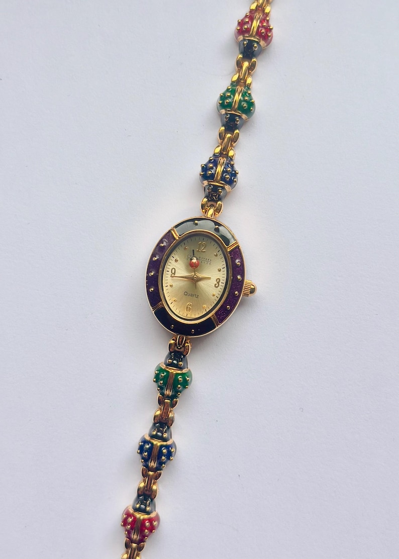 May include: Gold-toned quartz watch with an oval face and a gold band. The watch face has a gold background with black numbers and hands. The band features colorful, decorative beads in red, green, blue, and black.