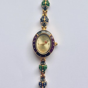 May include: Gold-toned quartz watch with an oval face and a decorative band. The watch face is gold with black numerals and hands. The band features colorful, jewel-like accents in red, green, and blue.