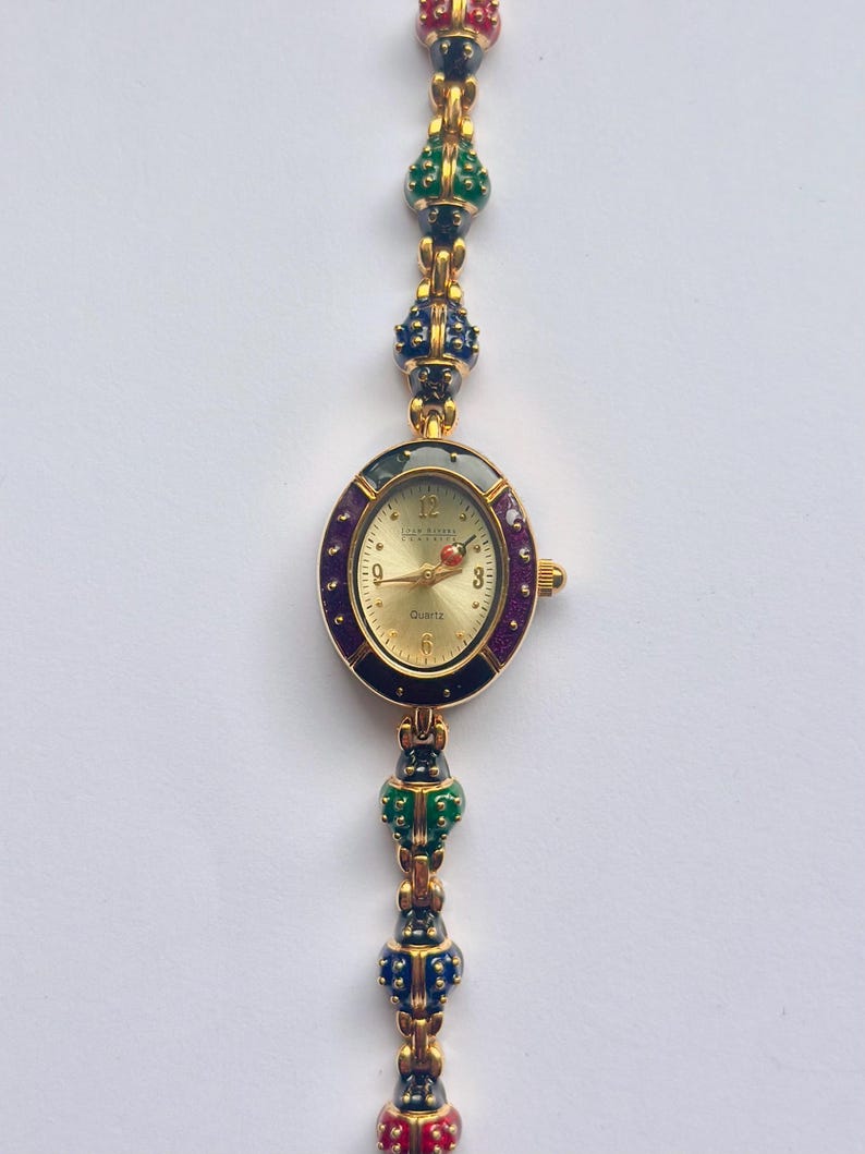 May include: Gold-toned quartz watch with an oval face and a decorative band. The watch face is gold with black numerals and hands. The band features colorful, jewel-like accents in red, green, and blue.