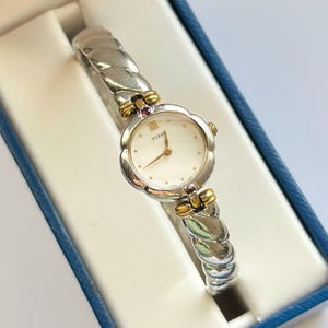 May include: A silver and gold-tone Tissot wristwatch with a white face and a decorative band. The watch is presented in a blue box. The watch face has gold-tone hands and hour markers. The band has a woven design.