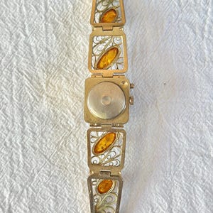 May include: A vintage gold-toned bracelet featuring rectangular filigree links with amber-colored oval stones. The bracelet has a watch-like clasp and a delicate, ornate design. The amber stones add a warm contrast to the gold metal.