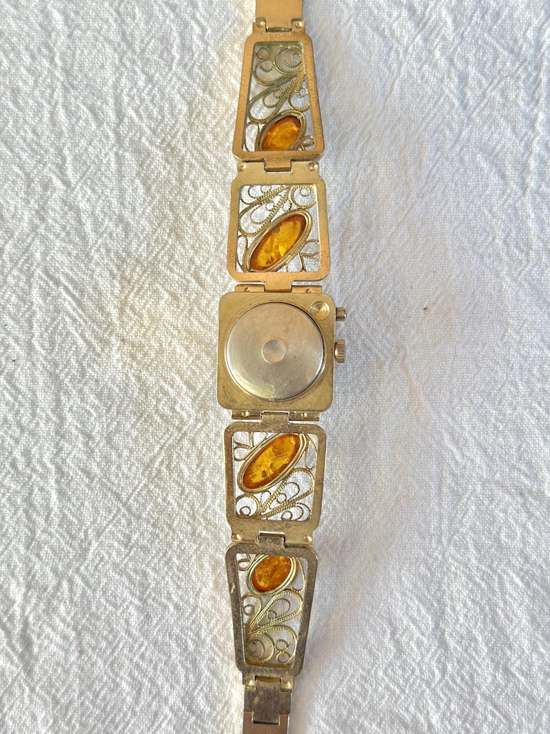May include: A vintage gold-toned bracelet featuring rectangular filigree links with amber-colored oval stones. The bracelet has a watch-like clasp and a delicate, ornate design. The amber stones add a warm contrast to the gold metal.