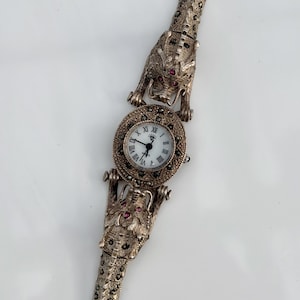 May include: A silver-toned watch featuring a round white dial with black Roman numerals and a diamond-shaped logo. The band and case are decorated with two dragon heads, each accented with small red stones and dark, textured metal detailing.