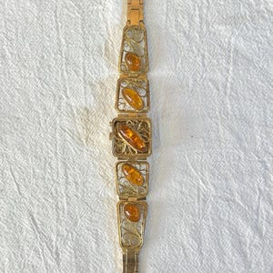 May include: A vintage gold-toned bracelet featuring rectangular filigree links, each holding an oval amber stone. The bracelet has a delicate, ornate design with a secure clasp. The amber stones have a warm, honey-like color, creating a striking contrast with the gold metal.