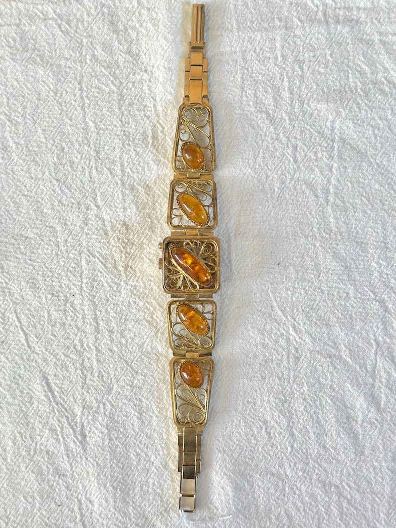 May include: A vintage gold-toned bracelet featuring rectangular filigree links, each holding an oval amber stone. The bracelet has a delicate, ornate design with a secure clasp. The amber stones have a warm, honey-like color, creating a striking contrast with the gold metal.